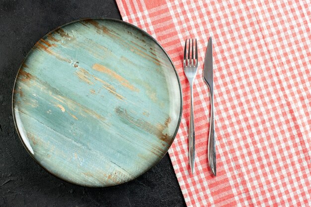 Top view round platter dinner knife and fork on red white checkered tablecloth on black table