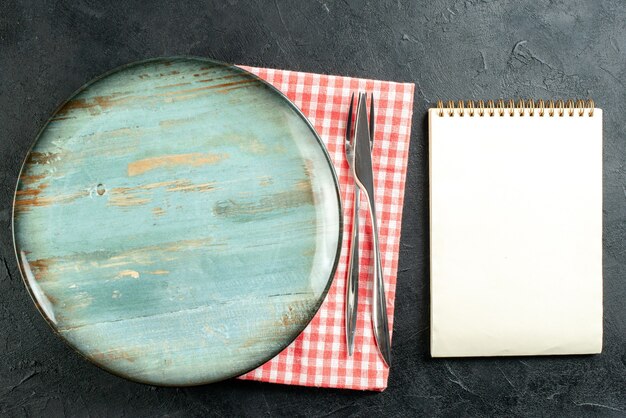 Top view round platter dinner knife and fork on red white napkin notepad on black table