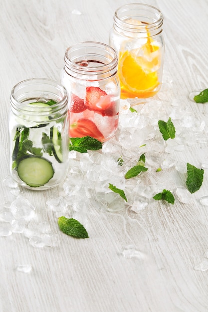 Free photo top view row rustic jars in crashed ice cubes with orange, strawberry, cucumber and mint prepared to make fresh homemade lemonade with sparkling water.