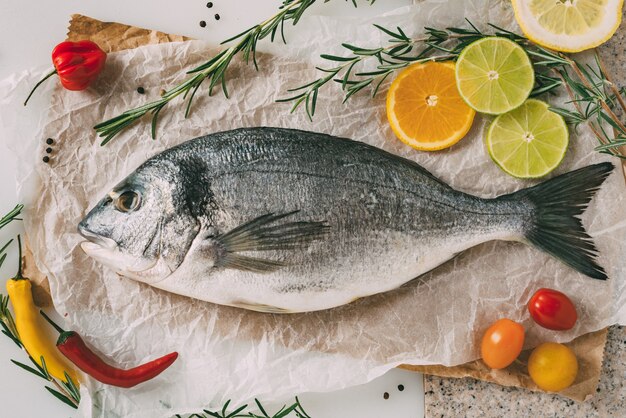 Top view of sea gilt-head bream fish on baking sheet with rosemary, lemon, orange, tomato