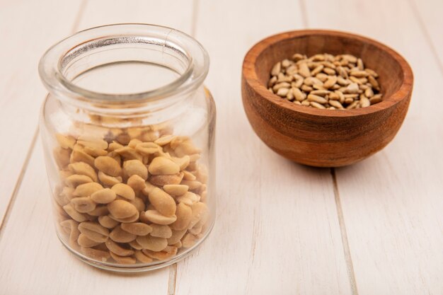 Top view of shelled sunflower seeds on a wooden bowl with pine nuts on a glass jar on a beige wooden table