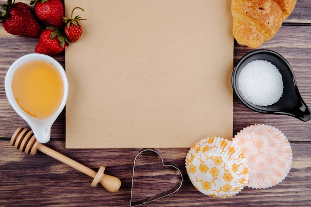 Top view of sketchbook and fresh ripe strawberries with honey sugar croissant and cookie cutters on wood