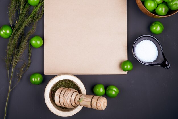 Top view of a sketchbook , salt , dried peppermint in a mortar , fennel and sour green plums scattered on black table