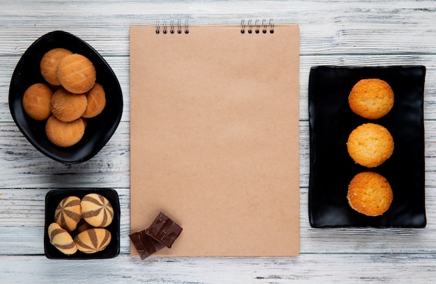 Top view of sketchbook and various types of sweet cookies on black trays on wooden background