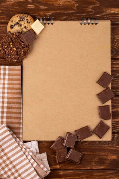 Top view of sketchbook with dark chocolate pieces and oatmeal cookies on wooden background