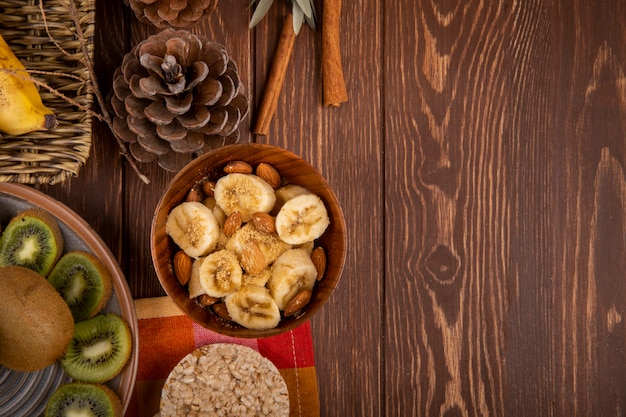 Top view of sliced bananas with almond in a wood bowl, slices of kiwi fruit on a plate and rice crackers on rustic with copy space