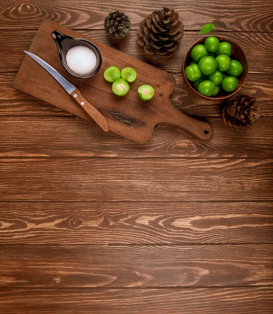 Top view of sliced green plums with salt and kitchen knife on a wooden cutting board , cones and plums in a bowl on rustic wooden table with copy space