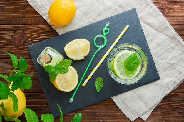 Top view sliced lemon in bowl with black cardboard, juice bottle, wooden knife and leaves on wooden surface. vertical