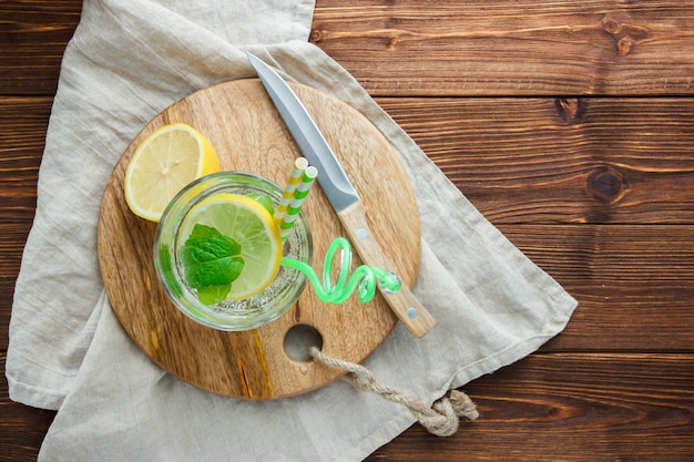 Free photo top view sliced lemon in bowl with cutting board, straw, white cloth, wooden knife on wooden surface. vertical