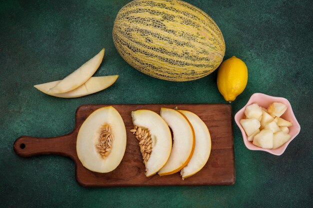 Top view of sliced melon on a wooden kitchen board with slices of melon on a pink bowl with lemon on green surface