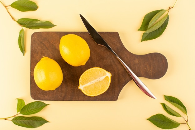 Free photo a top view sliced whole lemon fresh juicy mellow along with silver knife and green leaves on the brown wooden desk and cream background citrus orange