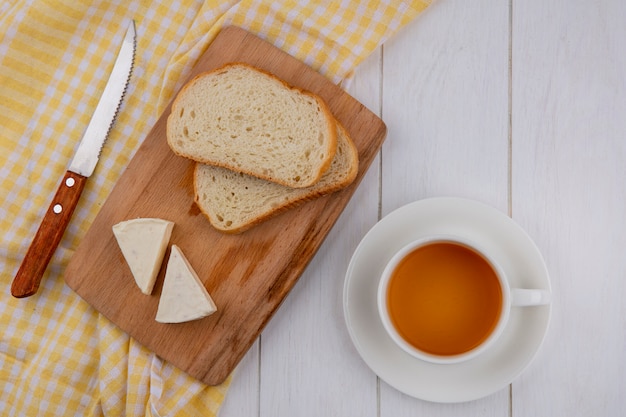 Top view of slices of bread with cheese on a blackboard with a knife on a yellow towel and a cup of tea on a white surface