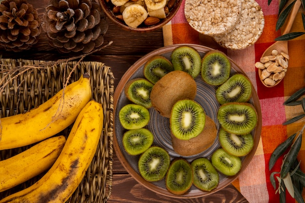 Free photo top view of slices of kiwi fruit on a plate and bunch of bananas in a wicker basket, wooden spoon with peanuts and rice crackers on wood