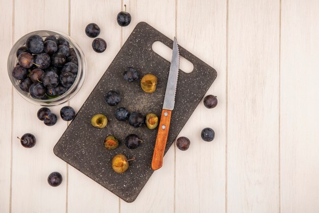 Top view of the small sour blue-black fruit sloes on a glass jar with slices of sloes on a kitchen cutting board with knife on a white wooden background