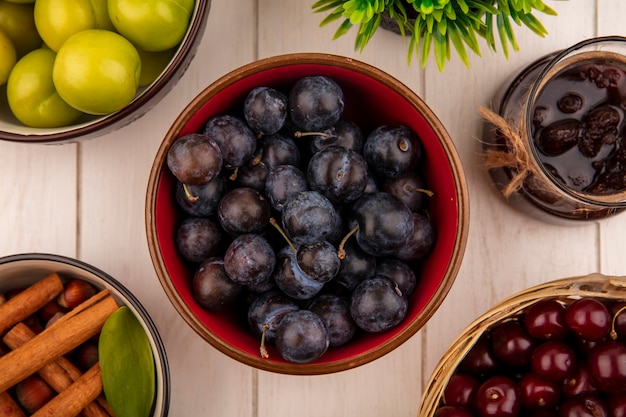 Free photo top view of the small sour blue-black fruit sloes on a red bowl with a strawberry jam with red cherries on a bucket on a white wooden background