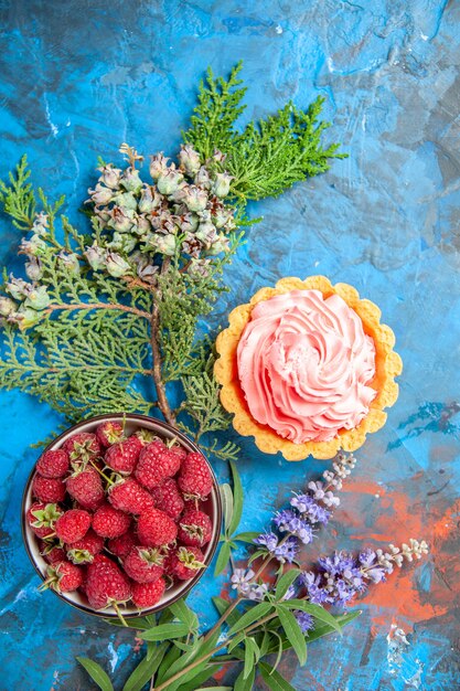 Top view of small tart with pink pastry cream bowl with raspberries on blue surface