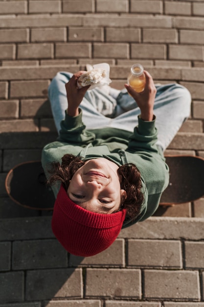 Free Photo top view of smiley teenager with skateboard eating a sandwich and drinking juice