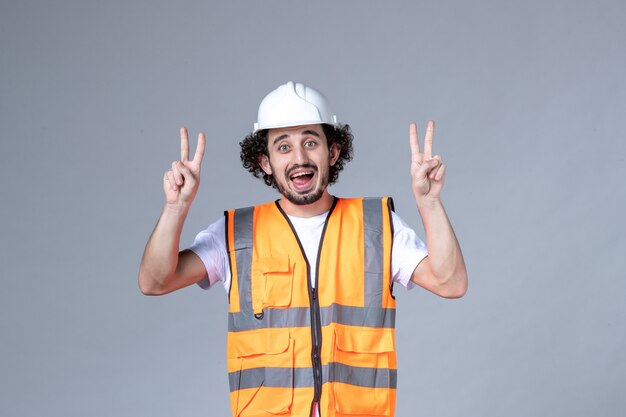 Top view of smiling male builder wearing warning vest with safety helmet and making victory gesture on gray wave wall