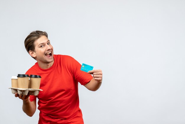 Top view of smiling young man in red blouse holding orders bank card on white wall