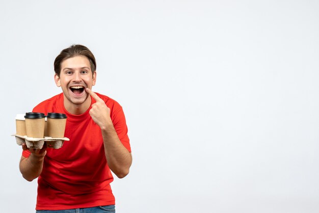 Top view of smiling young man in red blouse holding orders making smile gesture on white wall