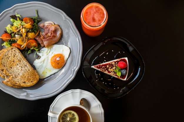 Top view of smoothie; cheesecake; tea; toast; salad; bacon; fried egg and toast on gray plate over black background