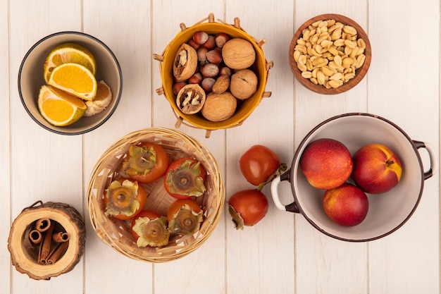 Top view of soft and juicy persimmons on a bucket with peaches on a bowl with nuts on a bucket with cinnamon sticks on a white wooden wall