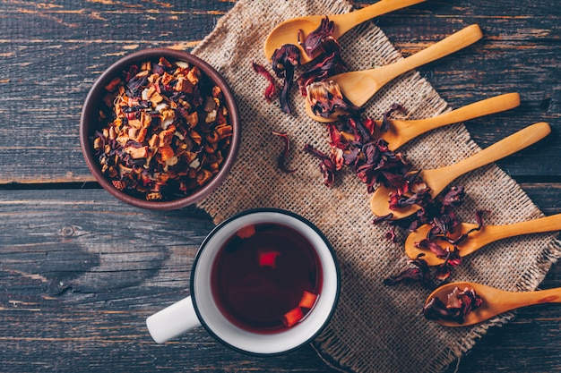 Free photo top view some kind of tea in bowl and spoons with a cup of tea on sack cloth and dark wooden background. horizontal