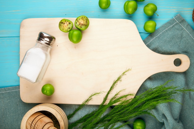 Top view of sour green plums and a salt shaker on a wooden cutting board with asparagus on blue table