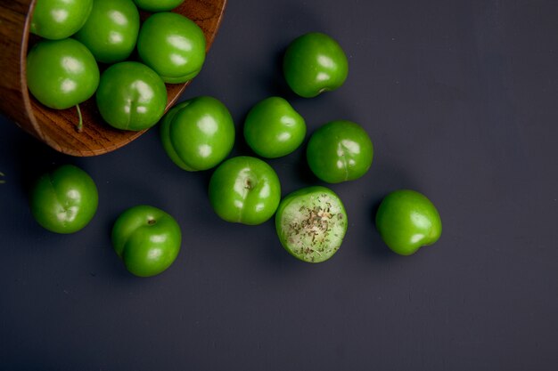 Top view of sour green plums scattered from a wooden bowl on black table