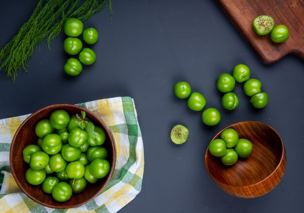 Top view of sour green plums in wooden bowls and wooden cutting board with sliced plums on black table