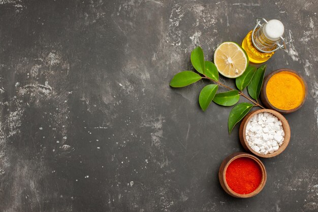 Top view spices bowls of colorful spices lemon with leaves next to the bottle of oil on the right side of the dark table