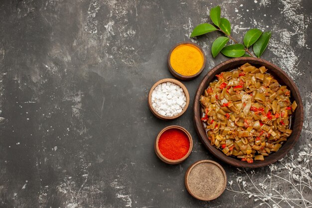 Top view spices plate of green beans next to the bowls of colorful spices leaves next to the tree branches on the dark table