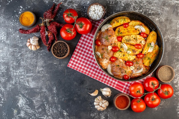 Top view of spicy raw chicken meal with potatoes vegetables in saucepan on red stripped towel and dried peppers garlics tomatoes yellow ginger on gray background