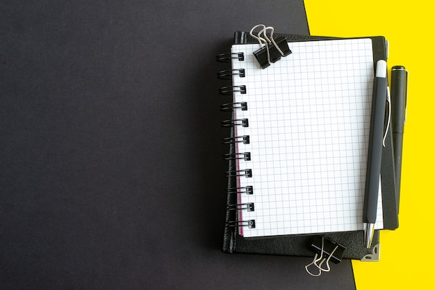 Top view of spiral notebook on book and pens on black yellow background with free space