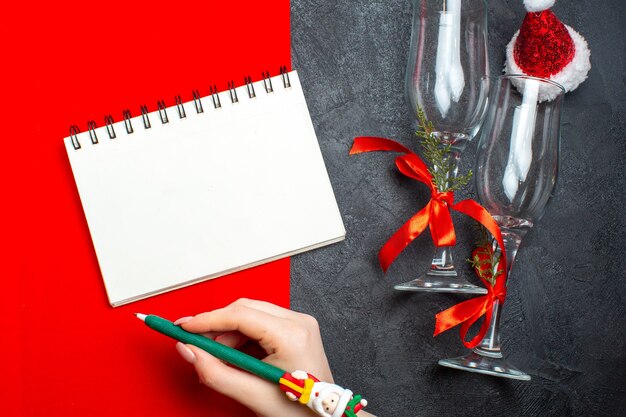 Top view of spiral notebook and hand holding a pen next to glass goblets santa claus hat on red and black background