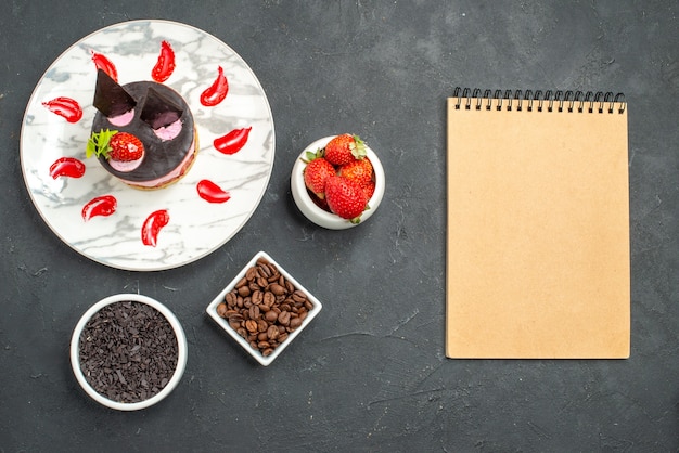 Top view strawberry cheesecake on white oval plate bowls with strawberries chocolate coffee bean seeds a notebook on dark surface