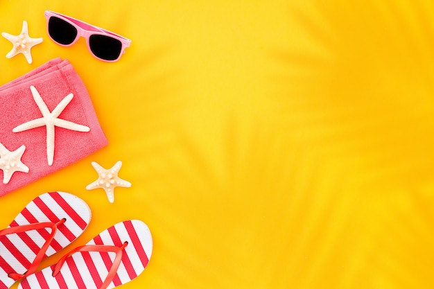 Top view sunglasses, towel starfish and flip flops, on yellow with sunlight and shadow of palm leaves. 