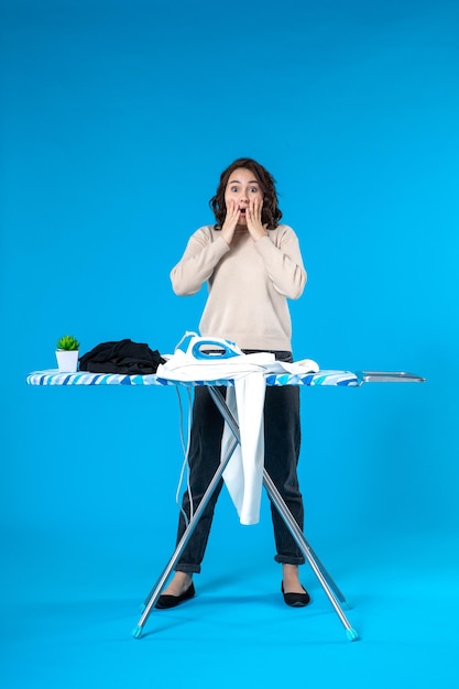 Top view of surprised young female standing behind the board and putting iron on clothes looking at it on blue surface