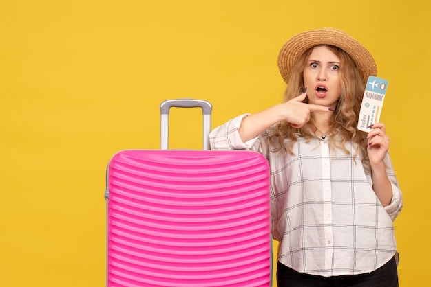 Top view of surprised young lady wearing hat pointing ticket and standing near her pink bag