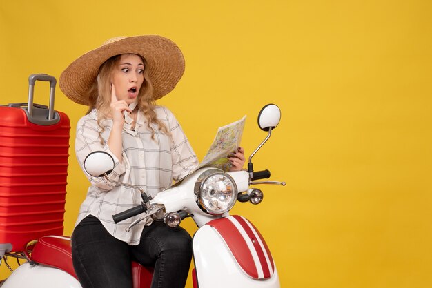 Top view of surprised young woman wearing hat and sitting on motorcycle and looking at map pointing up on yellow 