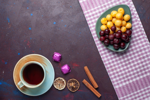 Top view of sweet cherries along with cinnamon and cup of tea on dark surface