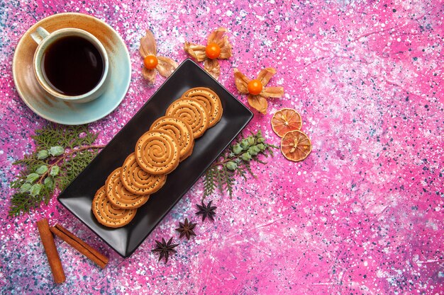 Top view of sweet cookies inside black form with cinnamon and tea on light pink surface