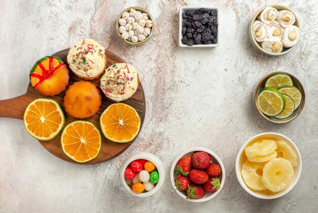 Top view sweets in bowls wooden board with cookies and orange next to the lemon strawberries dried pineapple and Turkish delight in bowls on the table