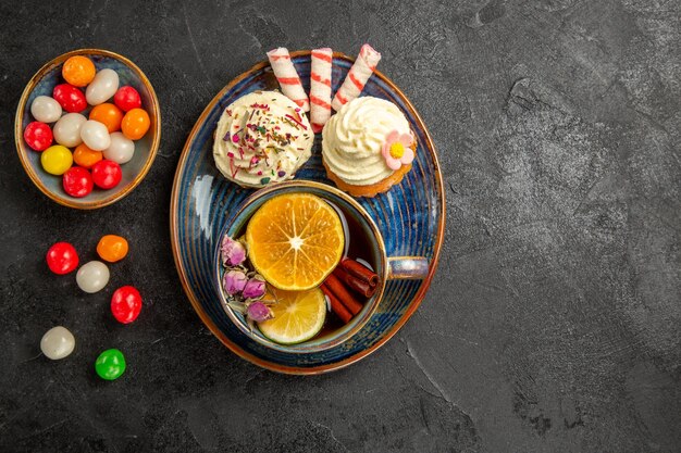 Top view sweets on the plate two appetizing cupcakes and sweets on the saucer a cup of tea with lemon and cinnamon sticks bowls of colorful sweets on the table