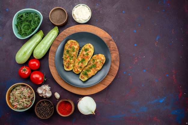 Top view of tasty baked squashes with greens along with fresh vegetables and seasonings on the dark surface