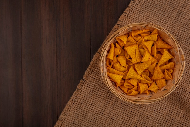 Top view of tasty cone shape fried corn snacks on a bucket on a sack cloth on a wooden table with copy space