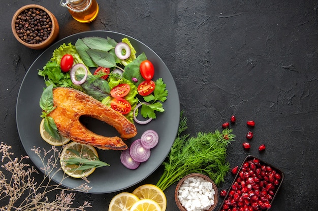 Top view tasty cooked fish with fresh vegetables and seasonings on dark table