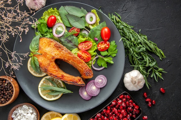 Top view tasty cooked fish with fresh vegetables and seasonings on dark table