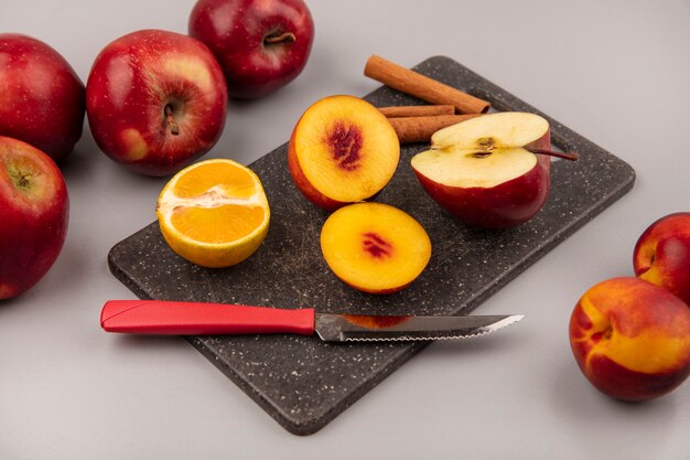 Top view of tasty half peaches on a black kitchen board with tangerine apple and cinnamon sticks with knife on a grey background