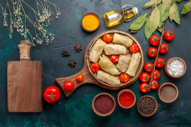 Top view tasty meat meal rolled inside cabbage with seasonings oil and fresh tomatoes on dark-blue desk